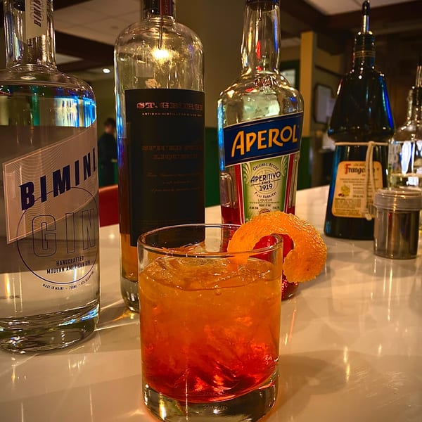 A vibrant orange cocktail with an orange peel garnish sits on a bar next to liquor bottles.