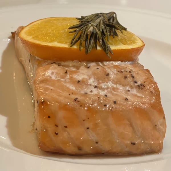 Close-up of a salmon fillet topped with an orange slice and rosemary sprig on a white plate.