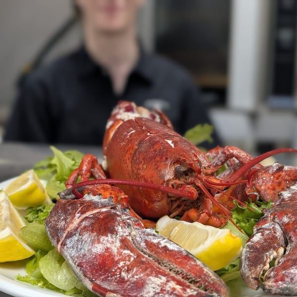 Whole cooked lobster on a bed of lettuce with lemon wedges, a server blurred in the background.