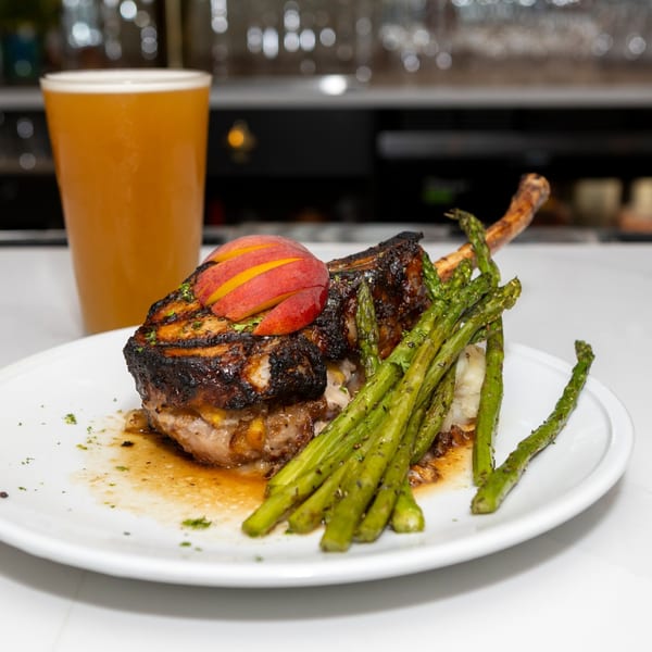 Grilled pork chop with sliced peaches, asparagus, and mashed potatoes, next to a glass of beer.